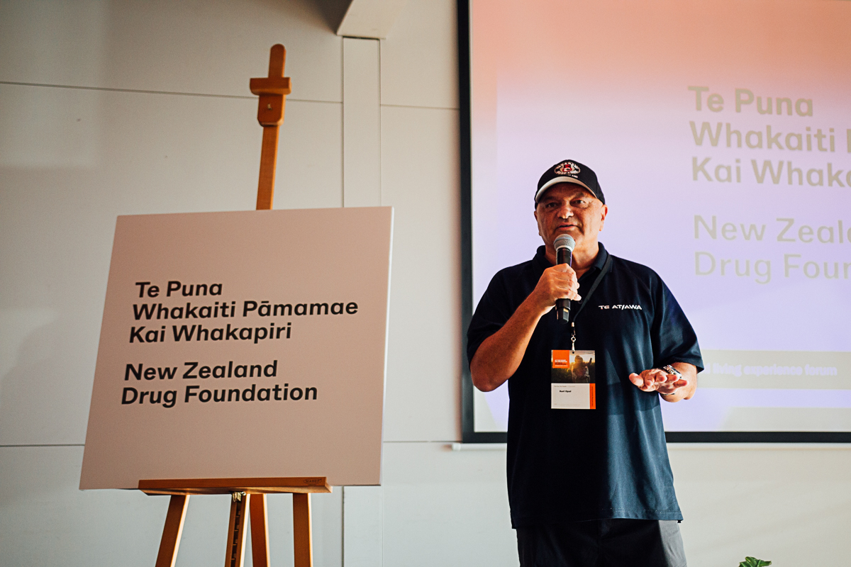 Māori man holding microphone, speaking next to sign that says "Te Puna Whakaiti Pāmamae Kai Whakapiri - New Zealand Drug Foundation"
